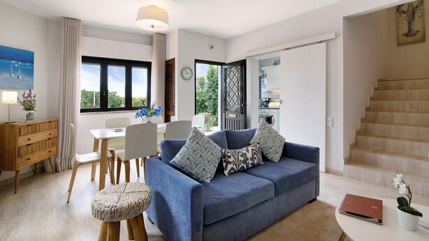A modern living room with a blue sofa, wooden dining table, and chairs. There is a staircase to the right and a door leading to another room. - Villa 'Harlequin' with Sea View, Wi-Fi and Air Conditioning (Vale do Lobo)
