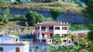 Un edificio rosa di due piani con un balcone, immerso nel verde e circondato da un pendio roccioso.