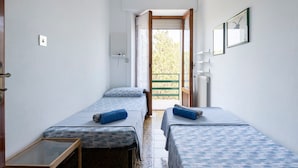 A small hotel room with two single beds, a wooden side table, a radiator, and a window with a view of greenery. - Apartment 'Oriente' with Private Terrace, Wi-Fi and Air Conditioning (Cala Gonone)
