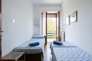 Quarto de hotel pequeno com duas camas de solteiro, uma mesa de cabeceira de madeira, um radiador e uma janela com vista para o verde.