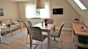 Uma sala de estar moderna com área de jantar, apresentando um sofá, uma mesa de centro, uma mesa de jantar com cadeiras, uma TV fixada na parede e uma janela com cortinas.