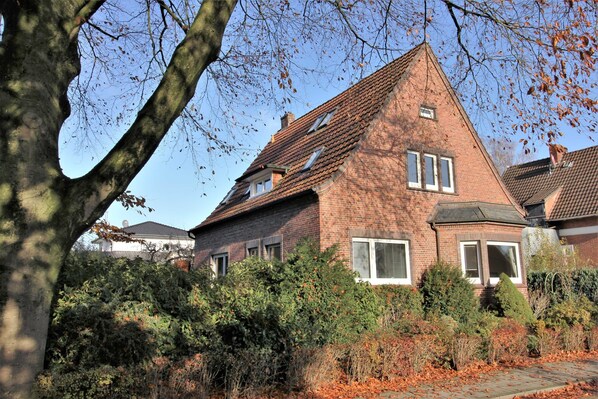 A brick house with a steeply pitched roof, surrounded by dense foliage and a leaf-covered ground.