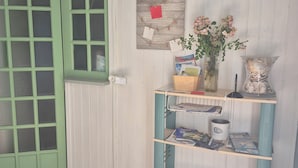 A room with a green door, a bulletin board with notes, a bookshelf with magazines, a vase with flowers, and a tiled floor. - Pleasant studio on the ground floor with private terrace and garden. 2 steps from the beach (Pornic)
