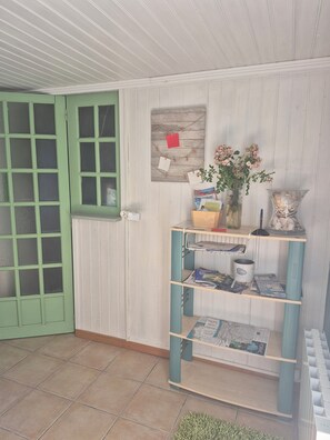 A room with a green door, a bulletin board with notes, a bookshelf with magazines, a vase with flowers, and a tiled floor.