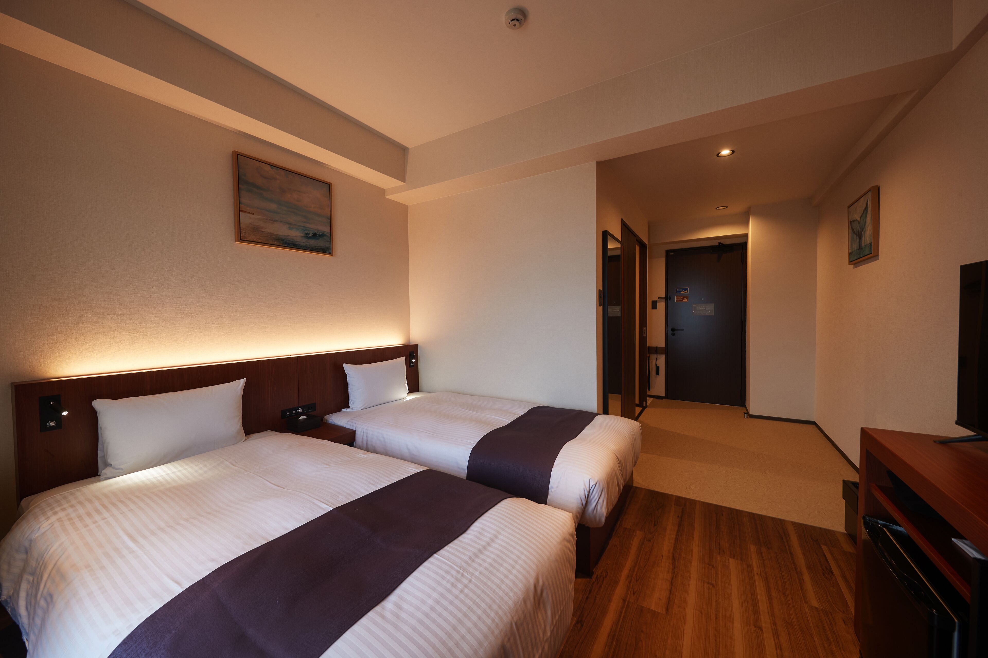 Quarto de hotel com duas camas, uma cabeceira de madeira, uma televisão de tela plana e um quadro na parede.