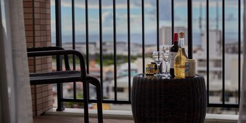 Un balcón con una mesa de mimbre que sostiene botellas y copas, una silla negra y vistas de la ciudad.