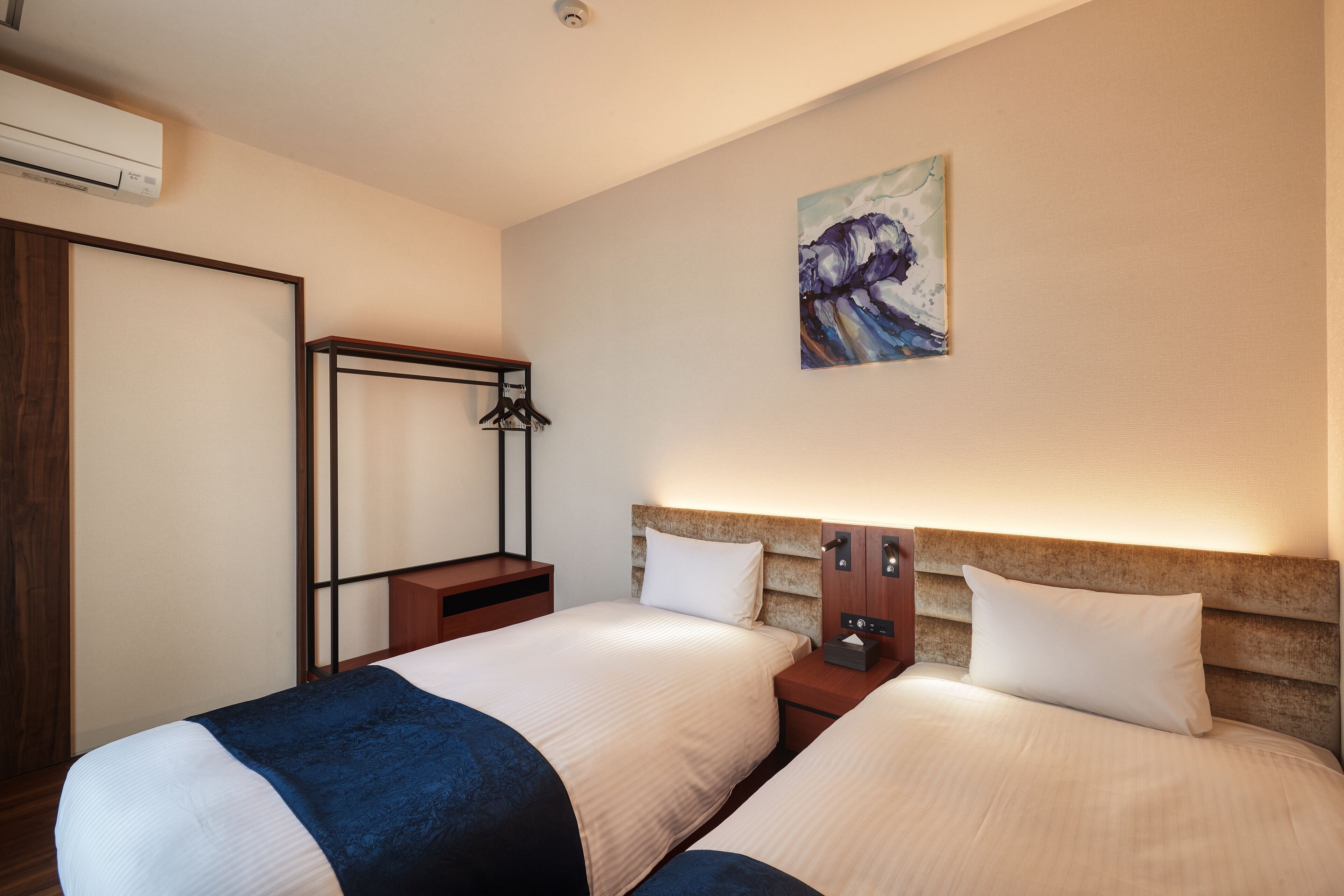 Une chambre d’hôtel avec deux lits, une armoire en bois, un miroir, un tableau accroché au mur et un appareil de climatisation.