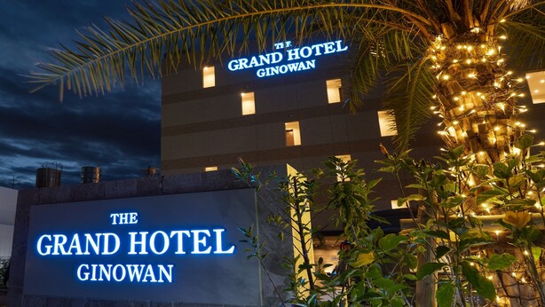 Exterior del Grand Hotel Ginowan con señalización iluminada, una palmera y fachada del edificio.