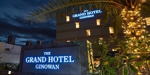 Exterior del Grand Hotel Ginowan con señalización iluminada, una palmera y fachada del edificio.