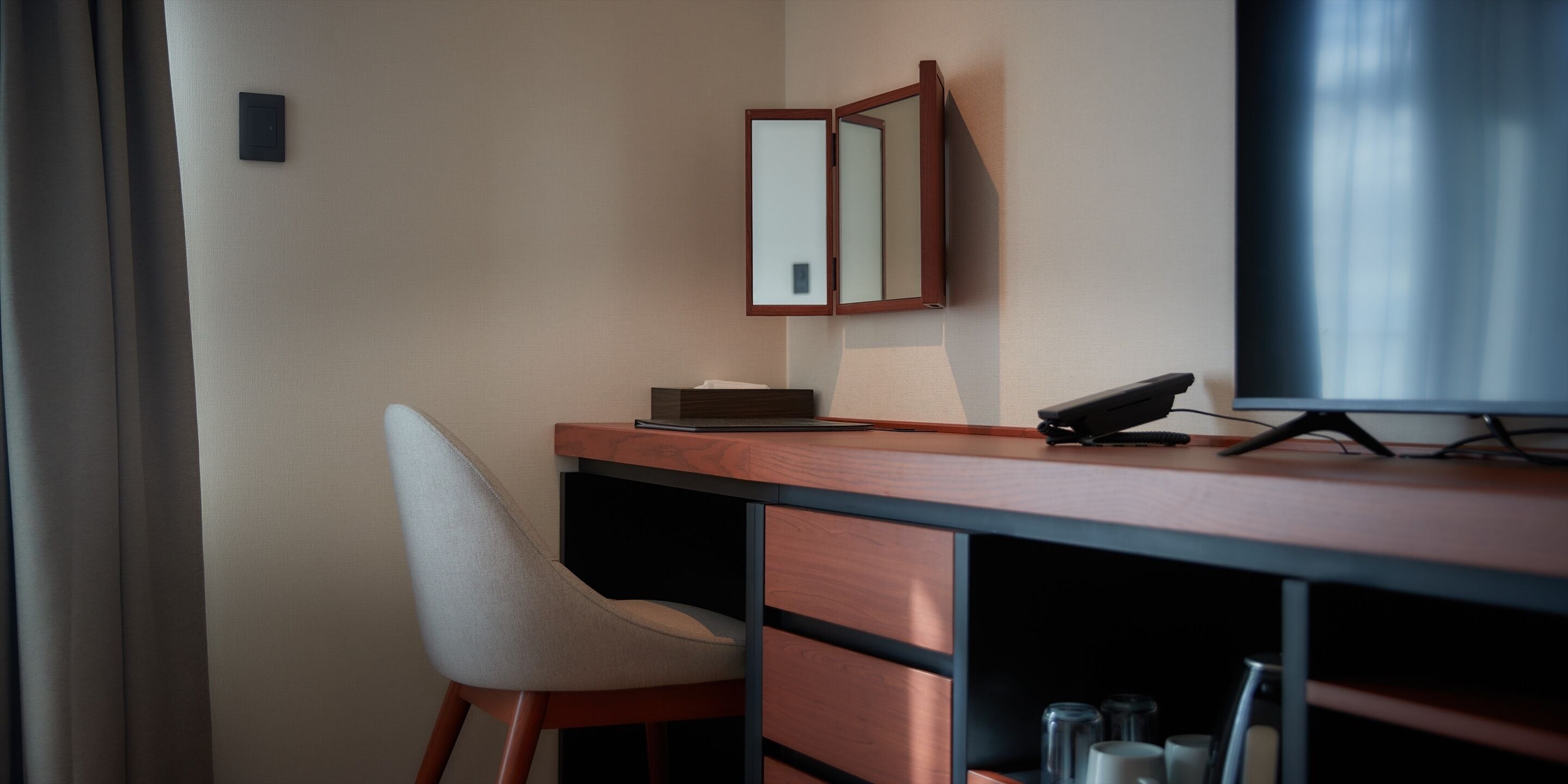 A hotel room with a wooden desk, a chair, a telephone, a mirror, and a television.