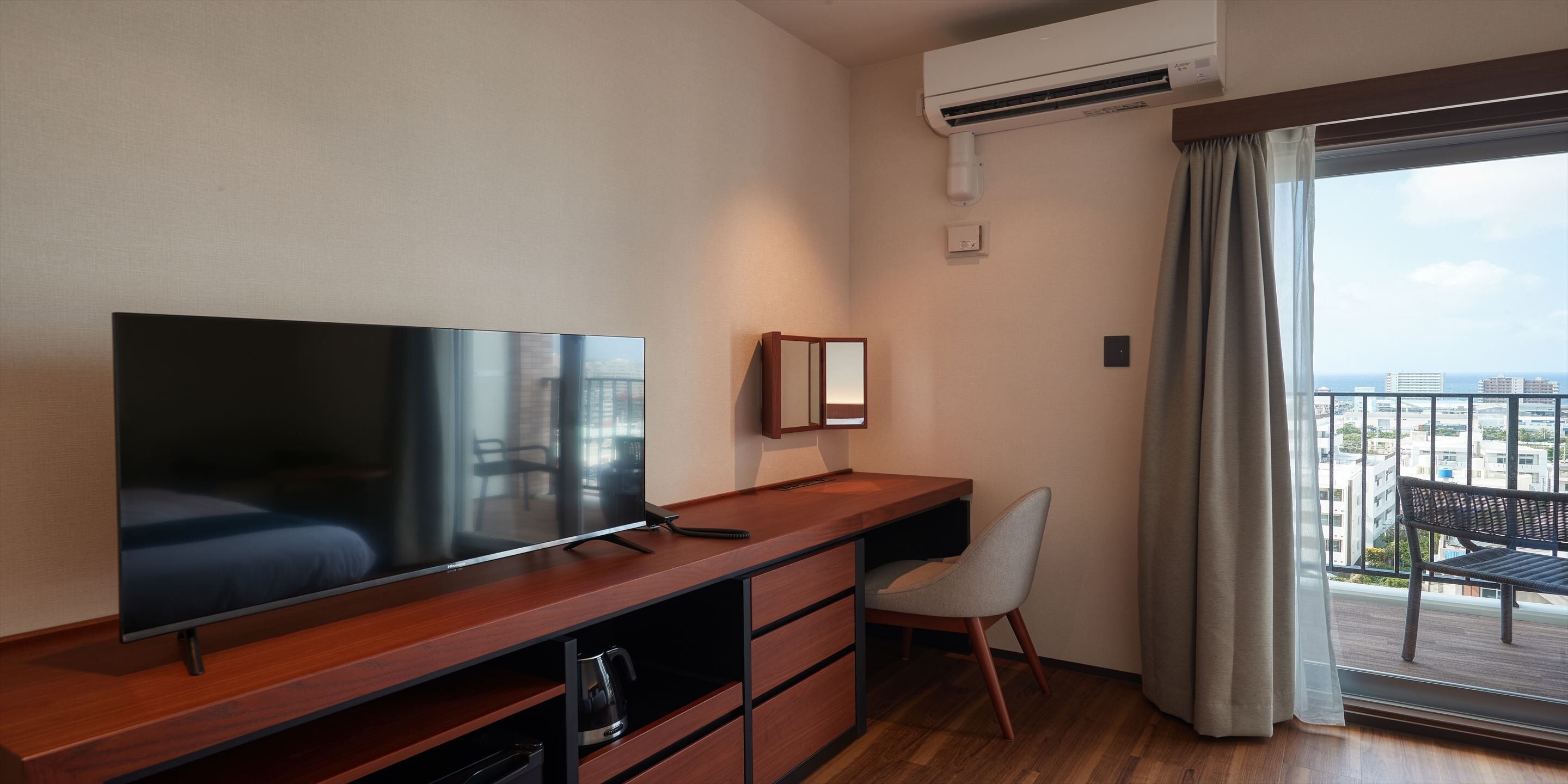 A modern hotel room with a large flat-screen TV, a wooden desk, a chair, and a balcony with a view of the city.