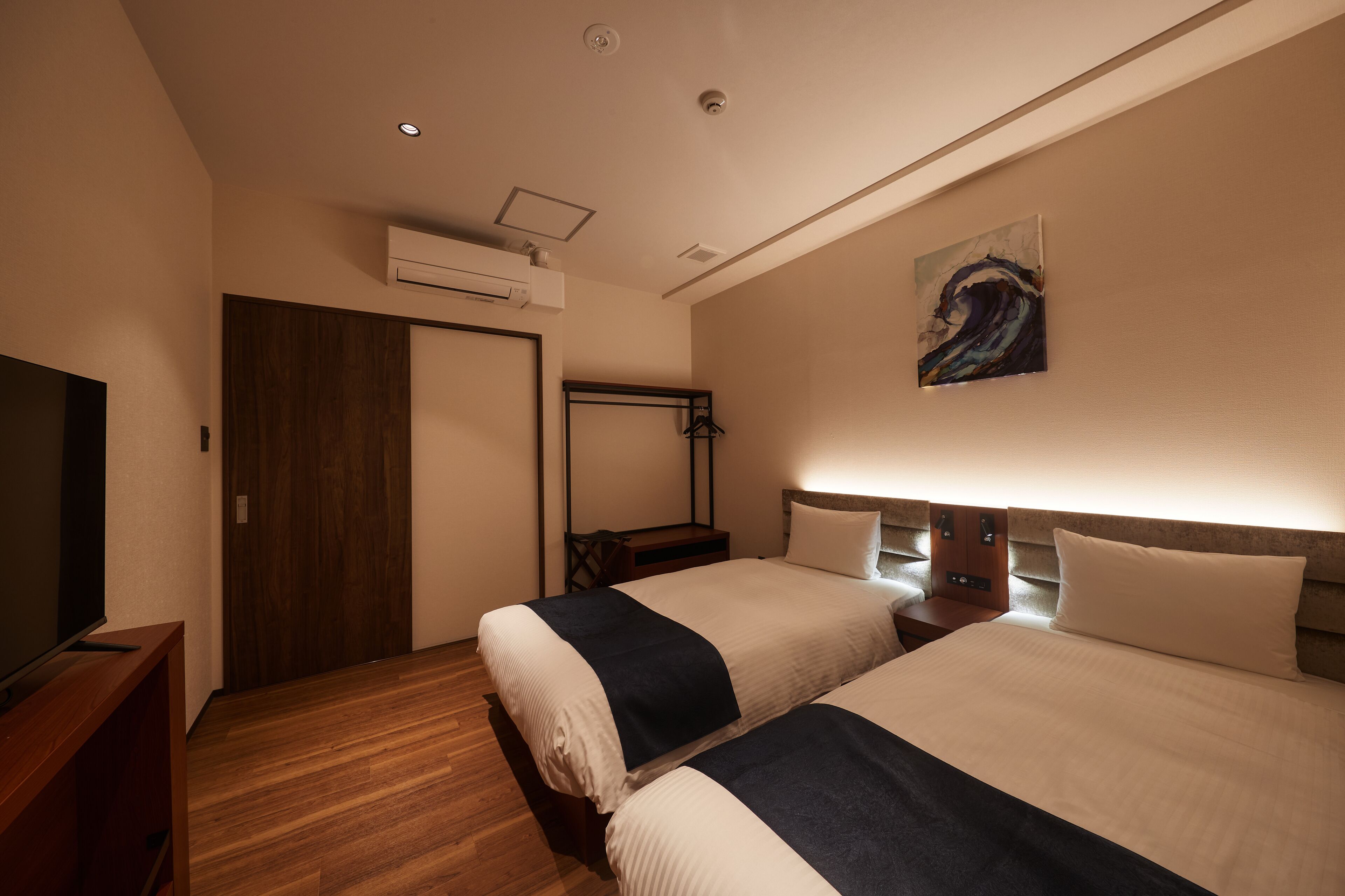 Habitación de hotel con dos camas, televisión, suelo de madera, aire acondicionado y un cuadro en la pared.