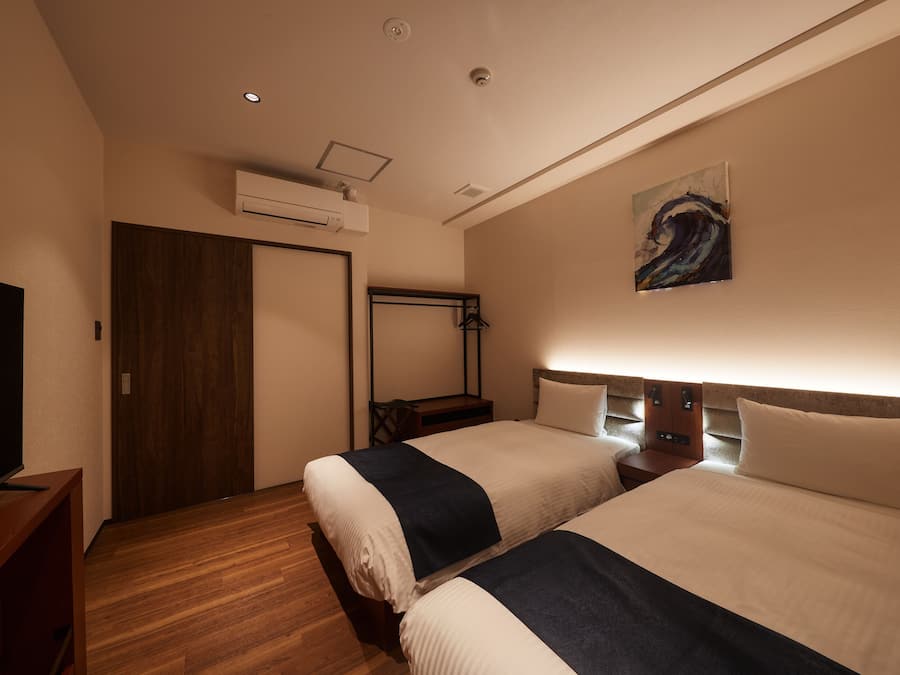 Habitación de hotel con dos camas, televisión, suelo de madera, aire acondicionado y un cuadro en la pared.