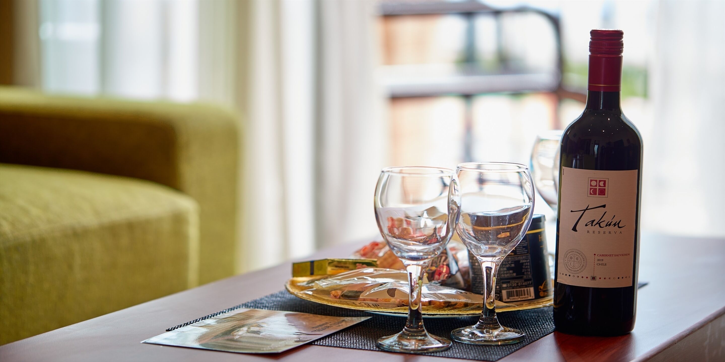 Une bouteille de vin Takun Reserve, deux verres à vin et un menu sont posés sur une table dans une pièce où se trouvent un canapé jaune et une fenêtre en arrière-plan.