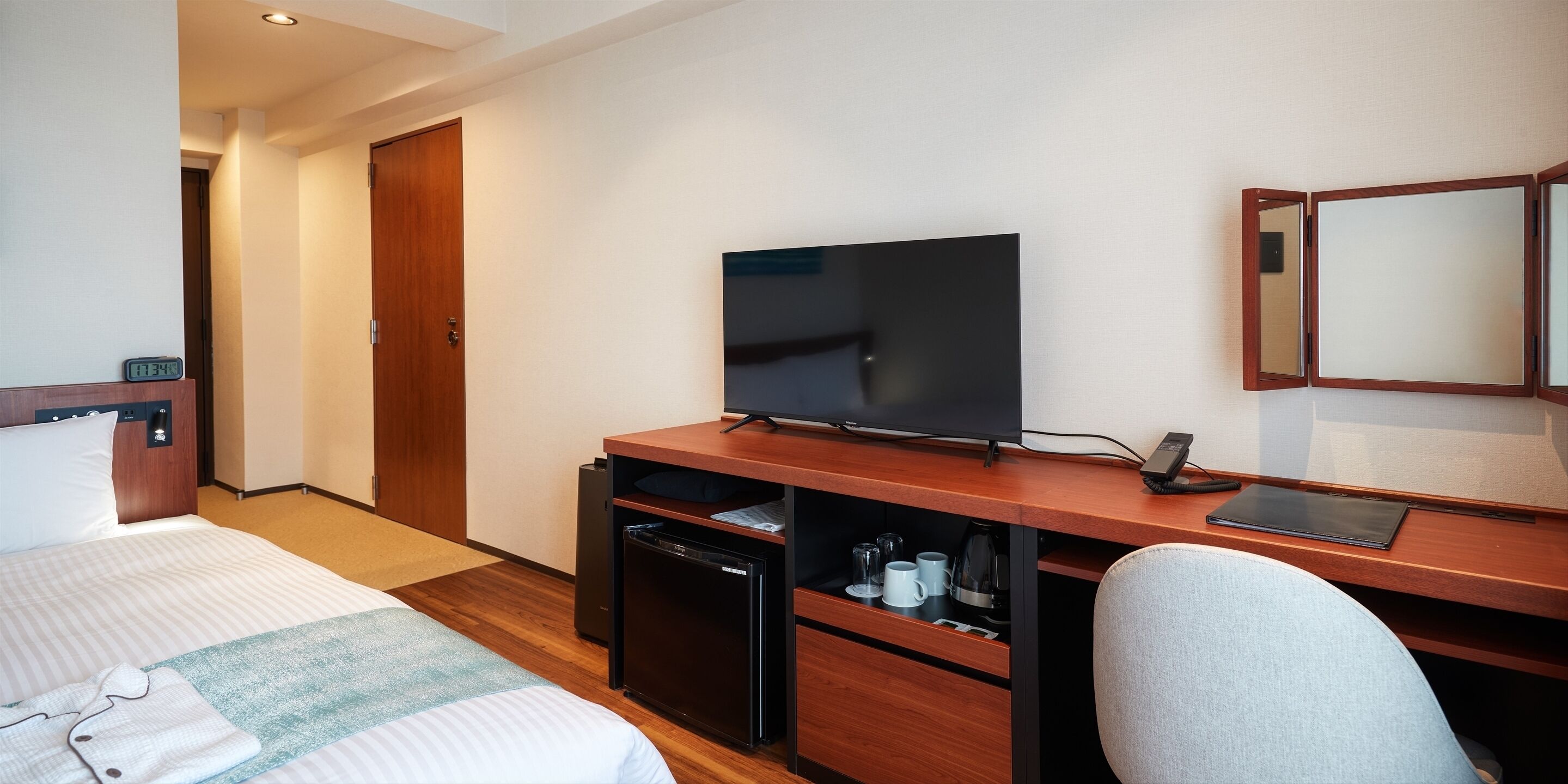 A hotel room with a bed, a desk with a television, a small refrigerator, and a mirror.