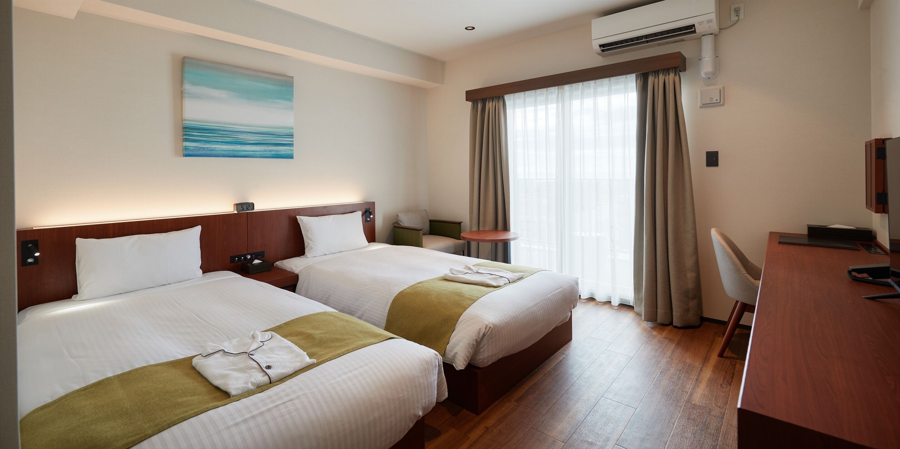 Habitación de hotel con dos camas, un escritorio, una silla, aire acondicionado y un cuadro en la pared.
