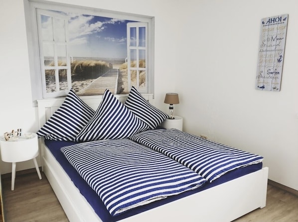 Uma cama com roupa de cama listrada, uma janela com vista para a praia, uma pequena mesa de cabeceira e um calendário de parede.