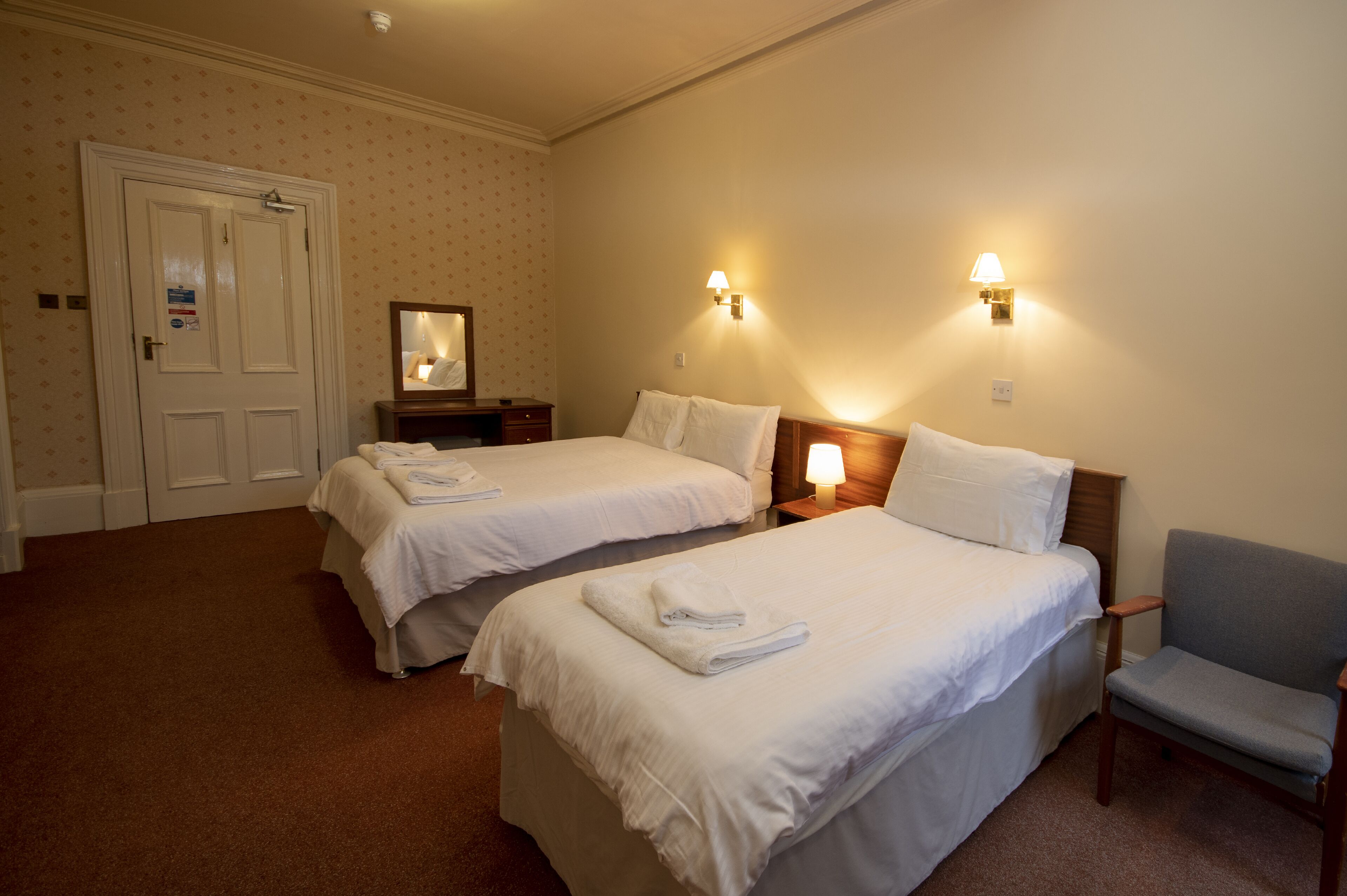A hotel room with two beds, a chair, a mirror, and two wall-mounted lamps.