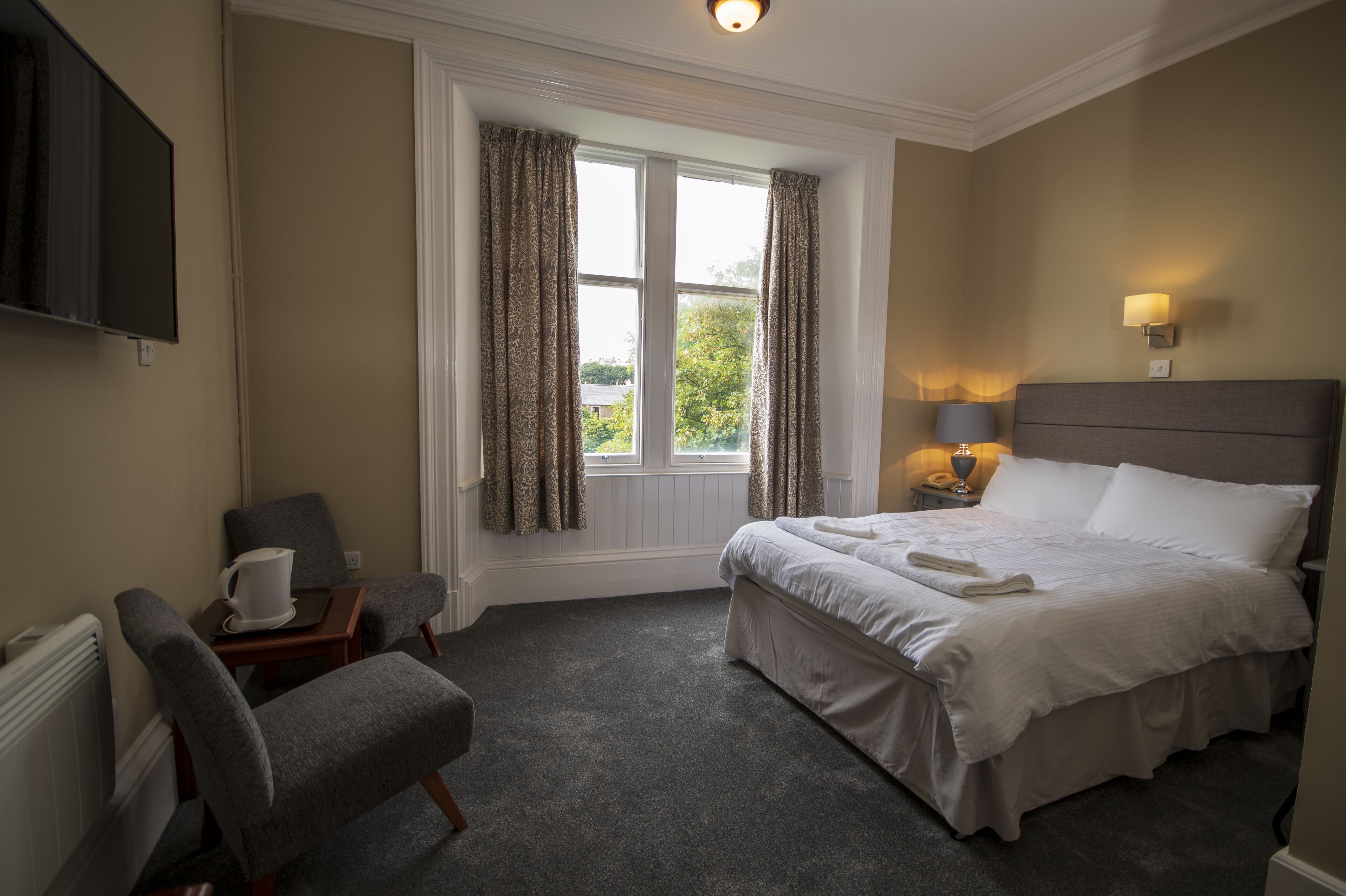 A hotel room with a bed, a chair, a table with a cup, a lamp, and a TV on the wall.