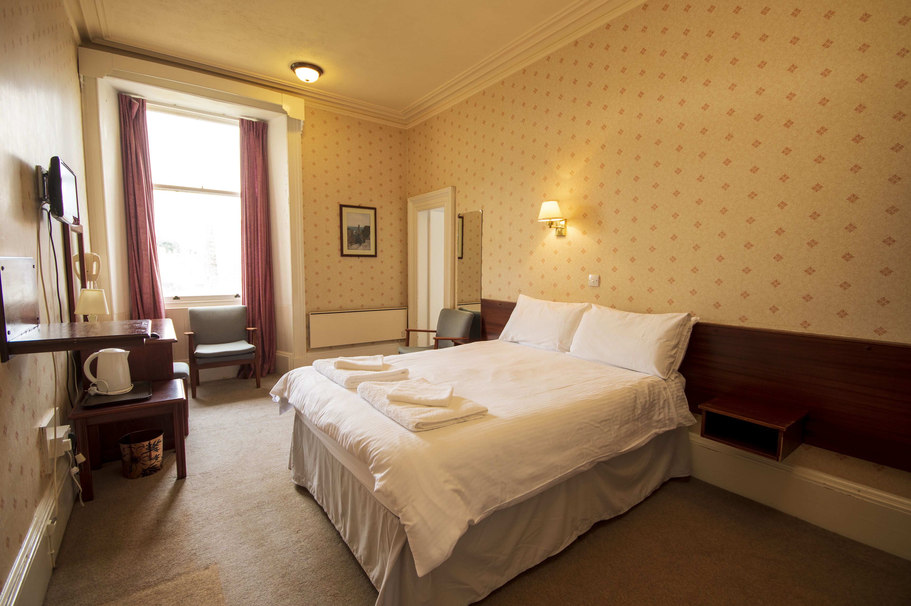 A neatly made bed with white linens, a bedside table with a lamp, and a chair in a hotel room with patterned wallpaper and a window with curtains.