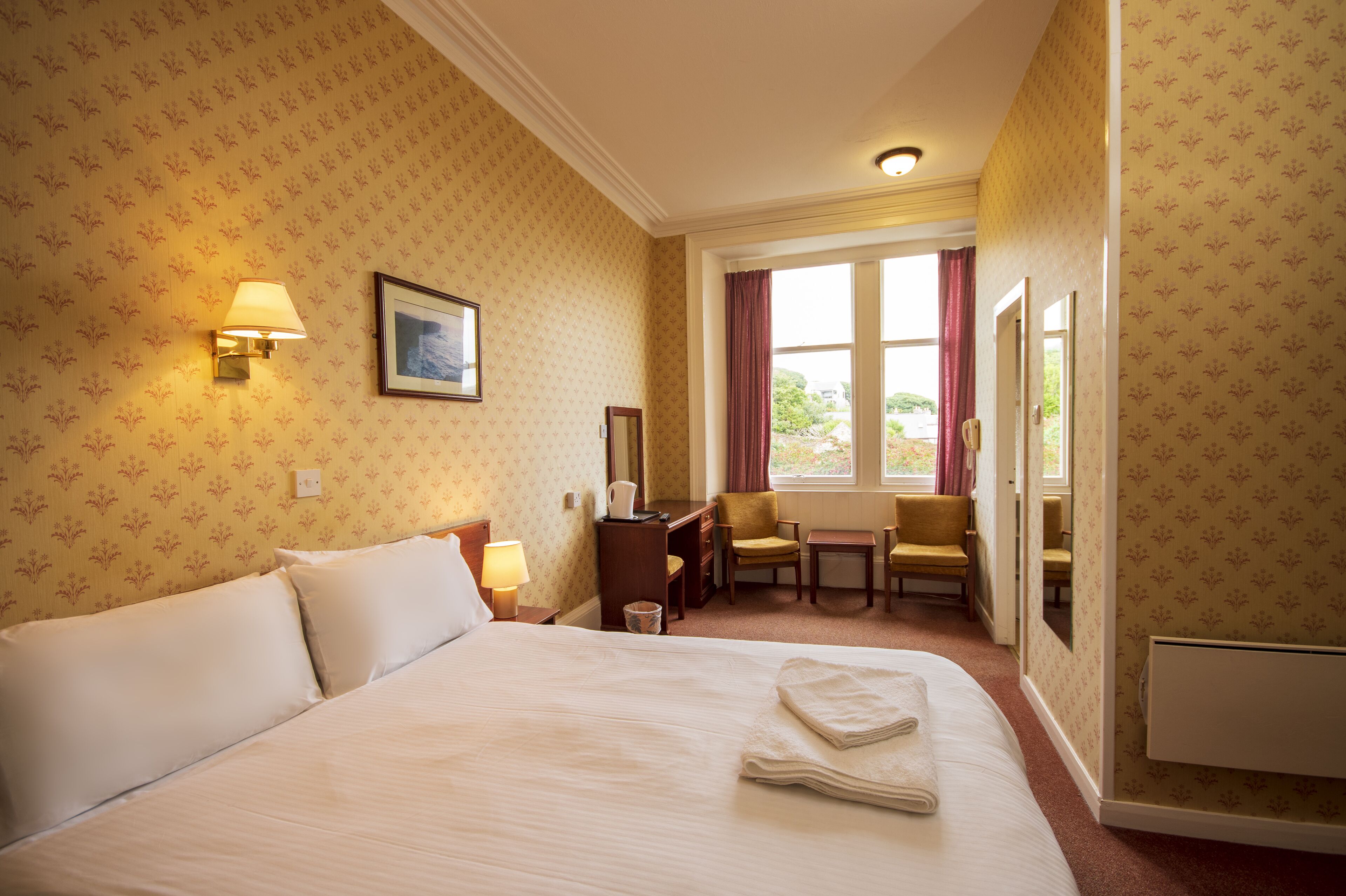 A hotel room with a large bed, a desk with a chair, a window with curtains, and a wall-mounted lamp.
