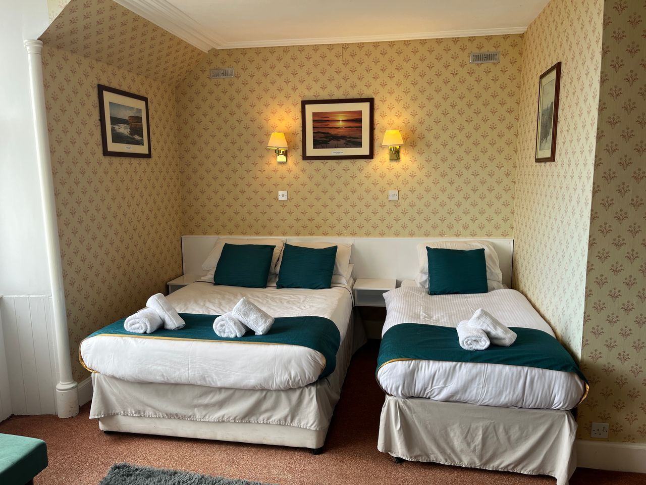 A hotel room with two beds, each with white linens and teal pillows, and two wall-mounted lamps.