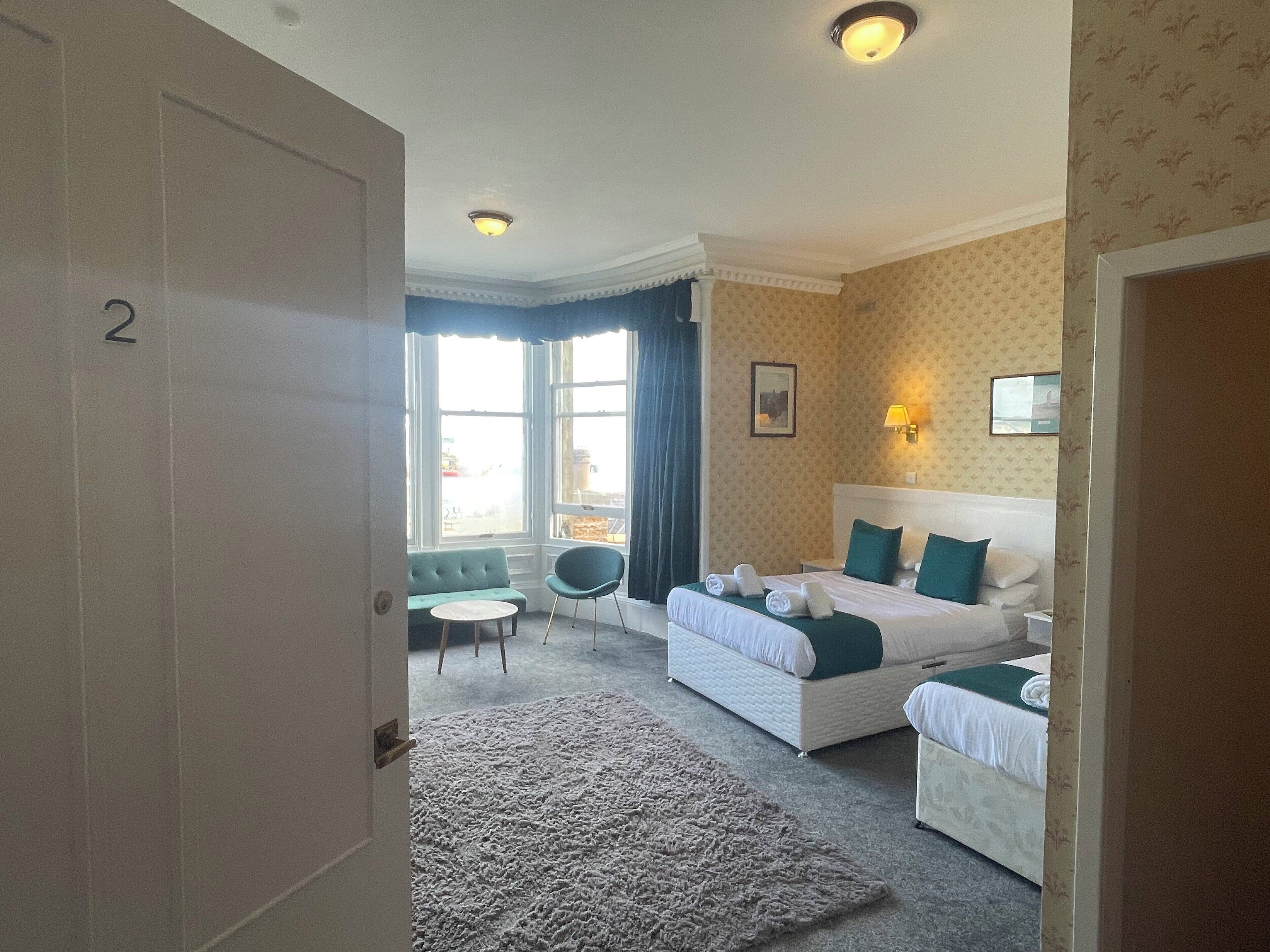 A hotel room with a large bed, a smaller bed, a sofa, a chair, and a view of the city through large windows.