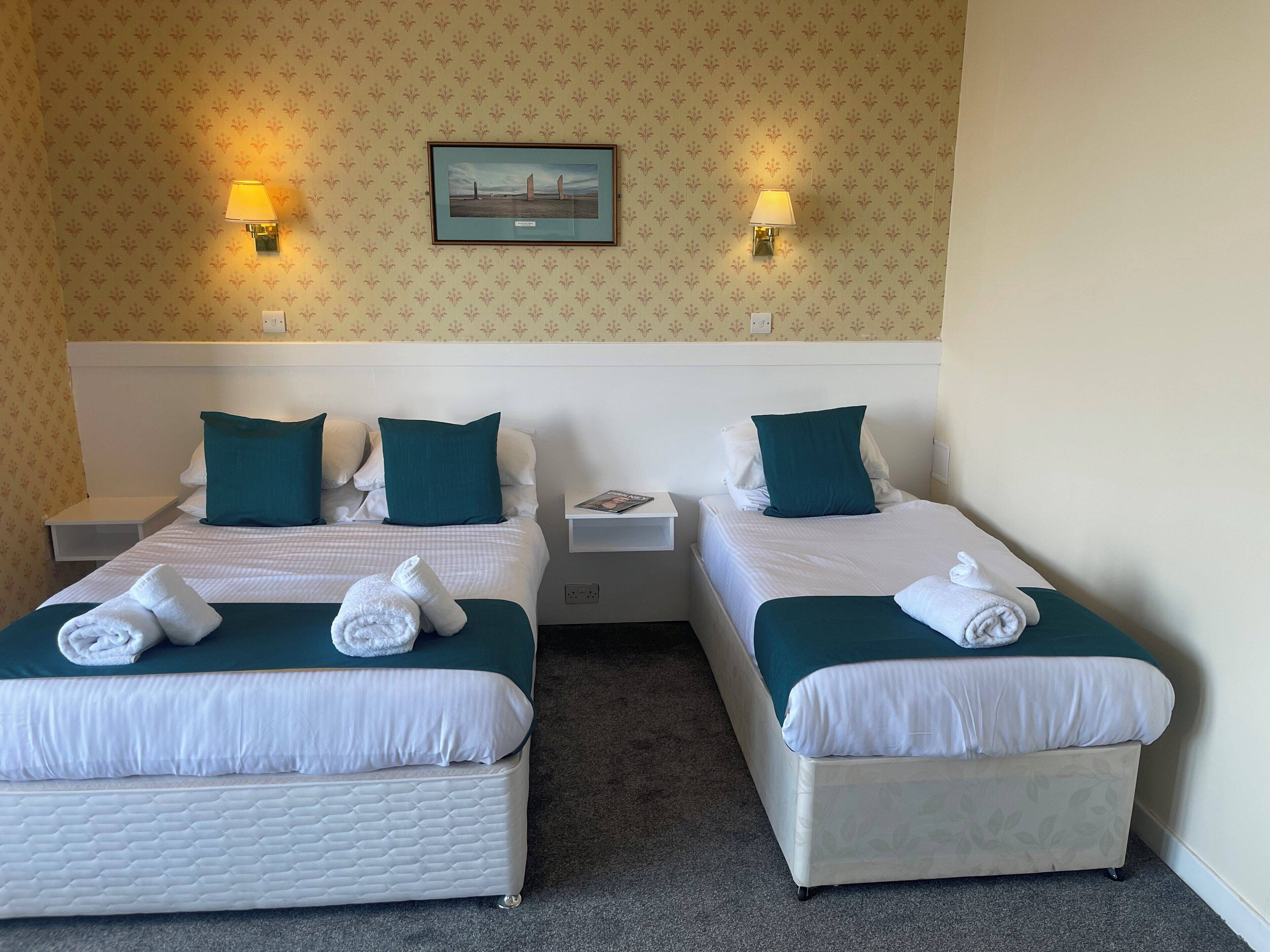 Habitación de hotel con dos camas, cada una con una almohada, una mesita de noche y una lámpara. Hay un cuadro enmarcado en la pared sobre las camas.