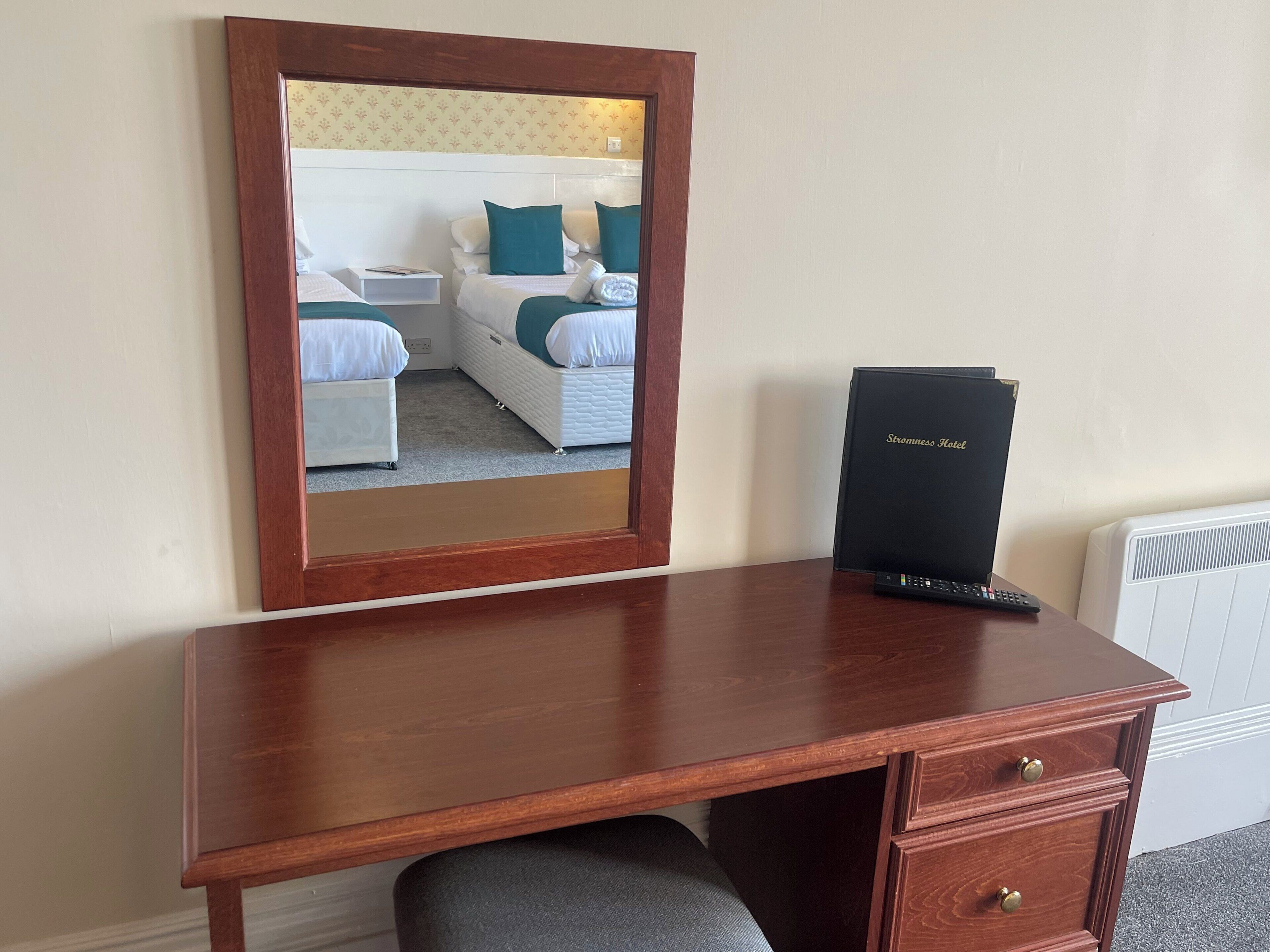 Een houten bureau met een spiegel die een hotelkamer weerspiegelt. De kamer heeft twee bedden met wit en teal beddengoed, een zwarte koffer en een zwarte doos met de tekst 'Storness Hotel'.