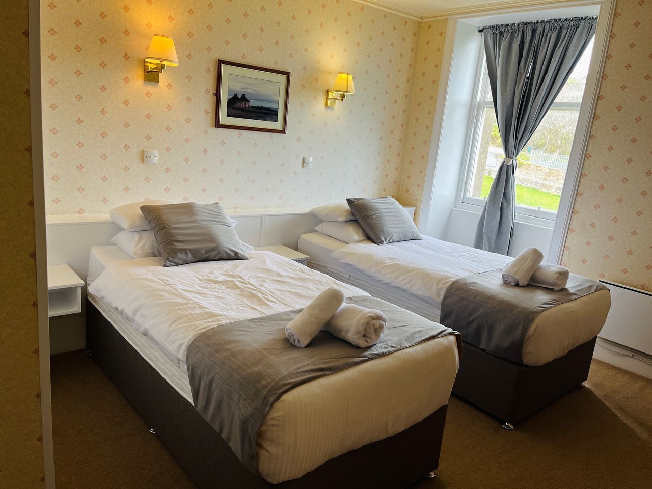 A hotel room with two beds, a nightstand, a lamp, a framed picture on the wall, and a window with curtains.