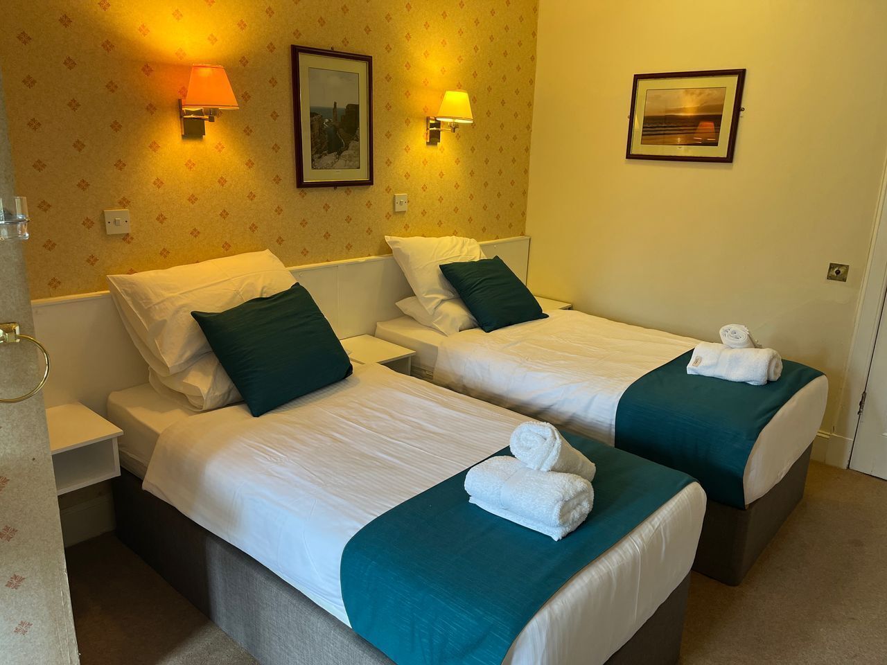 Habitación de hotel con dos camas, cada una con ropa blanca y colchas color turquesa. Hay dos lámparas de pared, un cuadro enmarcado y una puerta a la derecha.