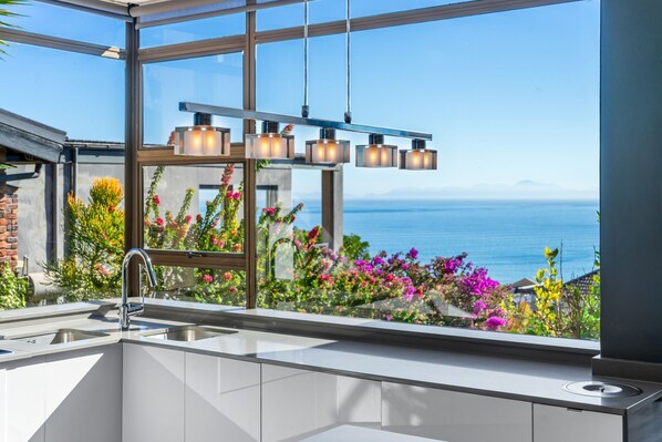 A modern kitchen with a large window overlooking a scenic ocean view, a built-in sink, and pendant lighting. - Zaria Sun Family (Cape Town)