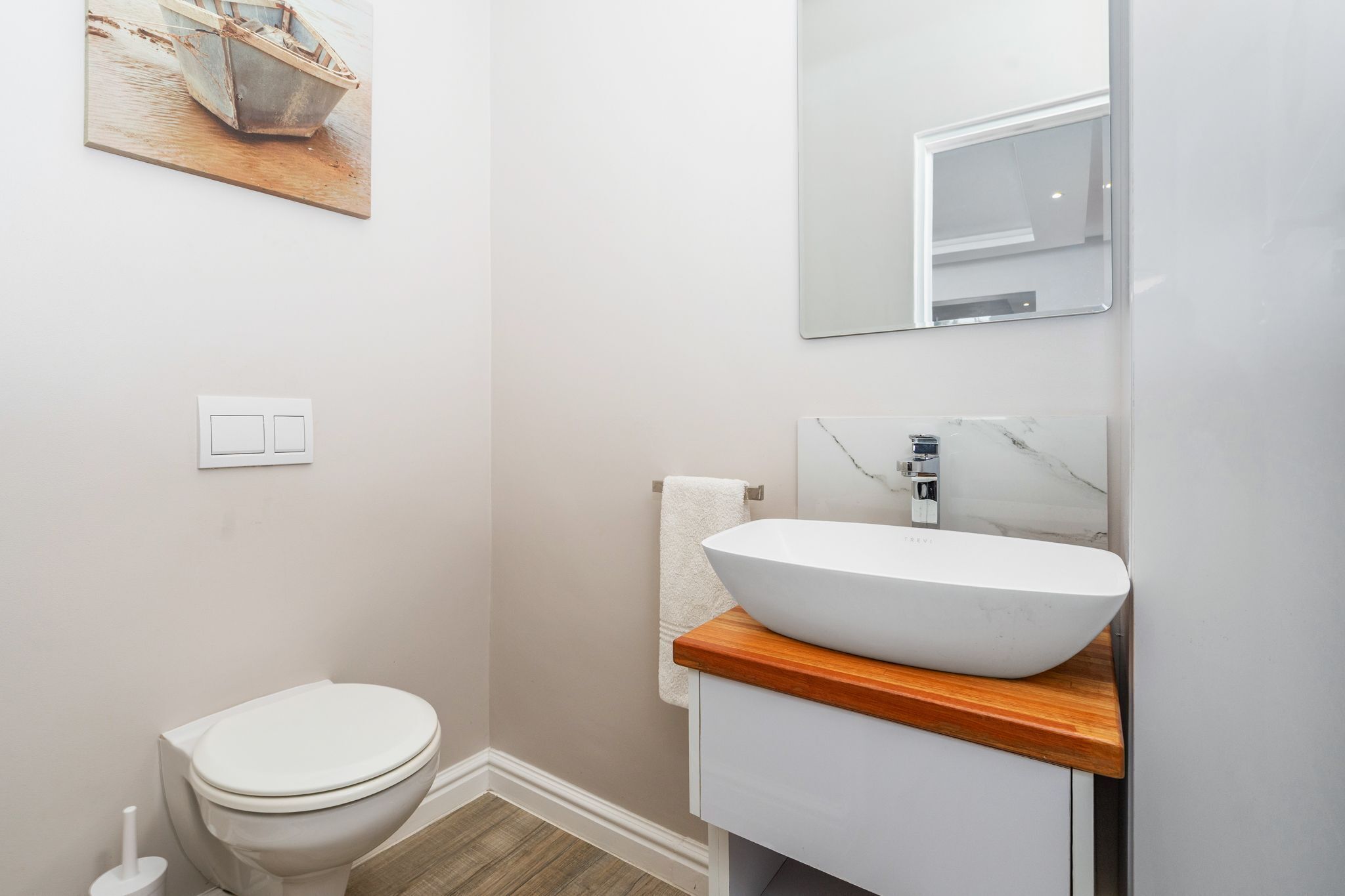 Een badkamer met een wit toilet, een houten wastafelmeubel met een witte wasbak en een schilderij van een boot aan de muur.