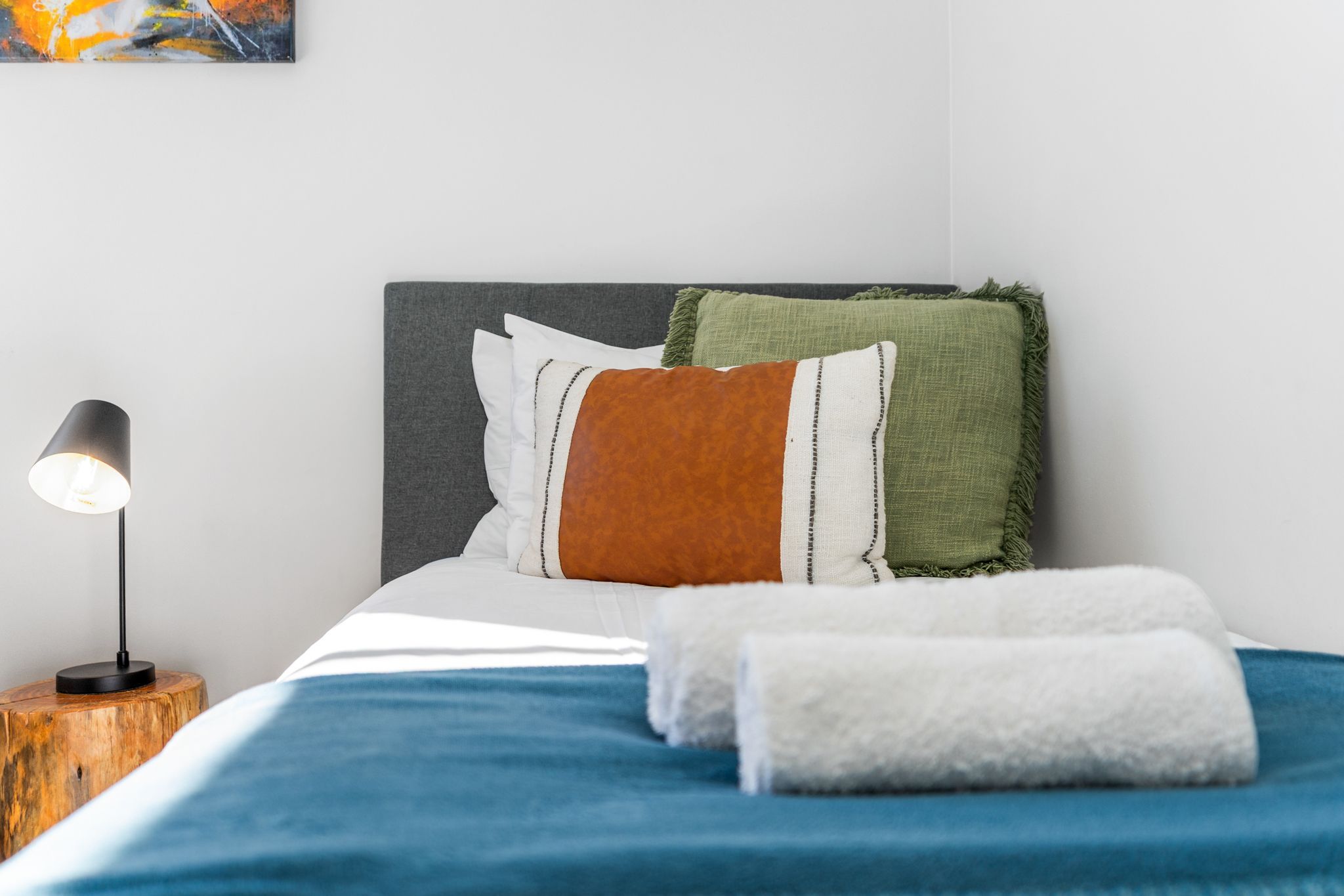 Ein ordentlich bezogenes Bett mit einer blauen Decke, einem weißen Kopfkissen und einem braunen Überwurf. Auf dem Kopfteil liegt eine grüne Decke. Am Fußende des Bettes ist ein weißes Handtuch gefaltet. Eine schwarze Stehlampe steht auf einem hölzernen Nachttisch neben dem Bett.