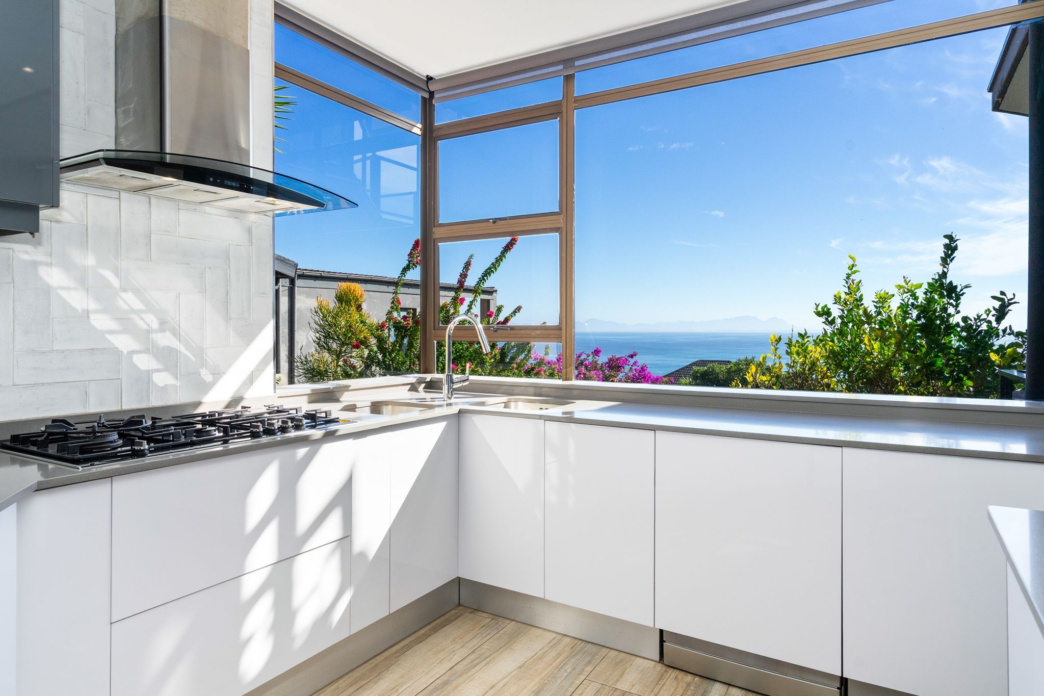 A modern kitchen with white cabinets, a stainless steel range, and a large window offering a view of the sea and mountains.