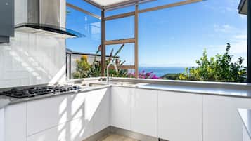 A modern kitchen with white cabinets, a stainless steel range, and a large window offering a view of the sea and mountains.