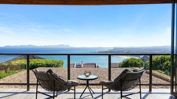 A balcony with two wicker chairs and a small table, overlooking a scenic view of the sea and distant mountains.