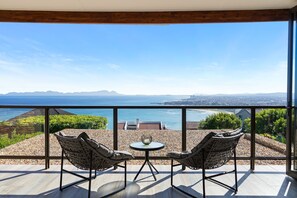 Een balkon met twee rieten stoelen en een kleine tafel, met uitzicht op een prachtig zeegezicht en verre bergen.