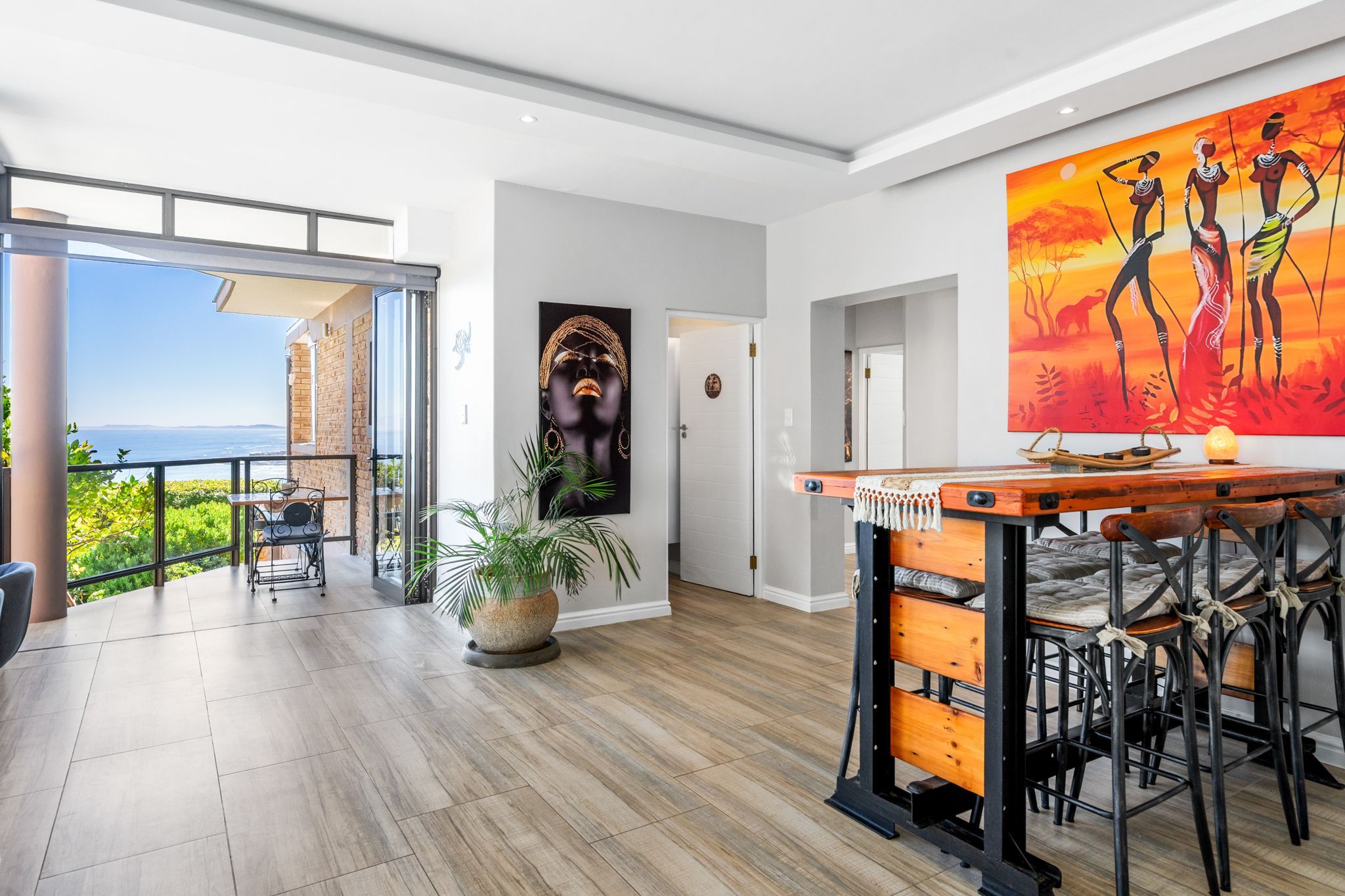 A spacious room with a large window offering a view of the sea, a dining area with a wooden table and chairs, and a vibrant painting on the wall.