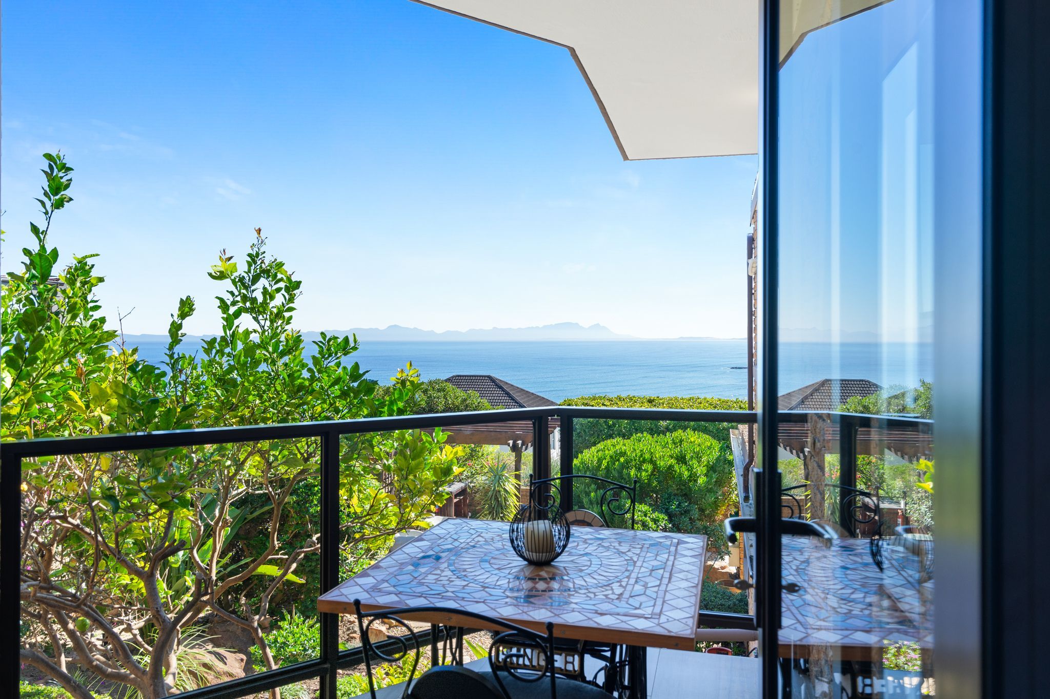 A balcony with a table and chairs, overlooking a scenic view of the sea and mountains.