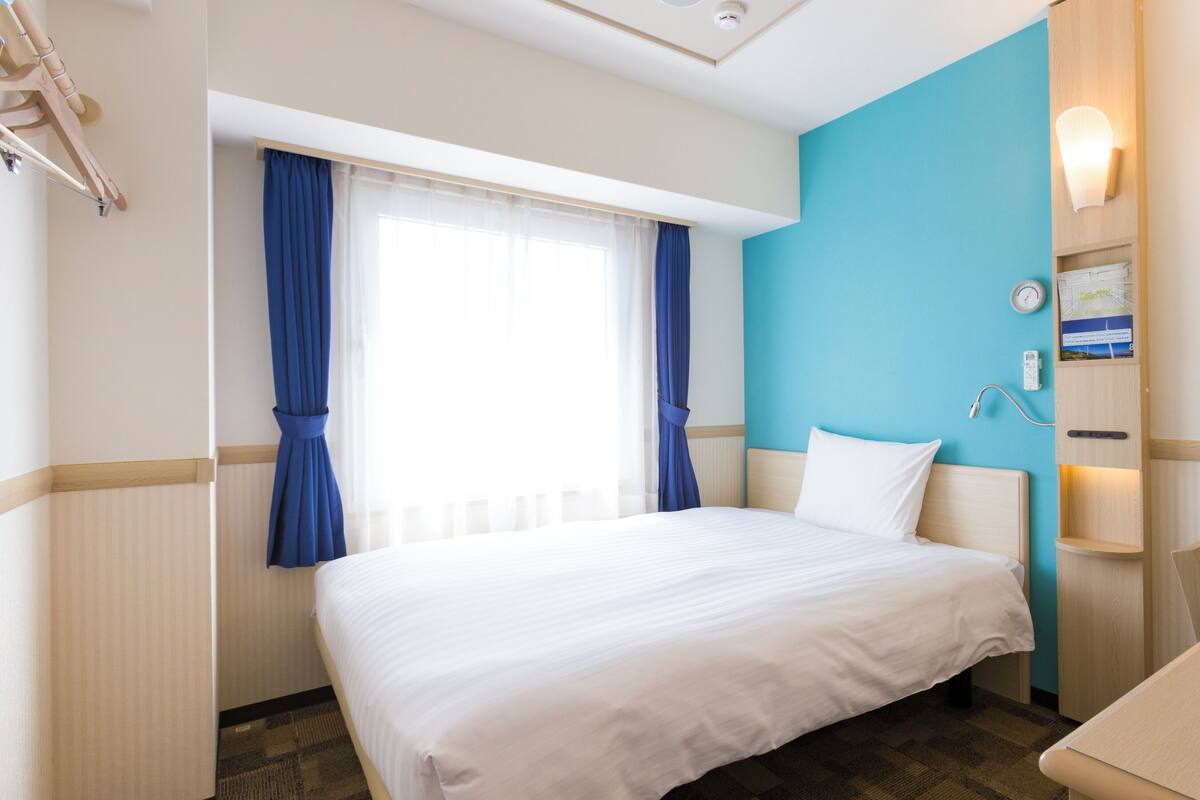 Een hotelkamer met een bed, een bureau, een stoel, een raam met gordijnen, een wandklok en een wandlamp.