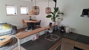 A modern kitchen with a central island, a dining area with a round table and chairs, a sofa with a yellow cushion, and a large potted plant. - 'Het Melkhuis', the tiny house that makes you happy😀 (Katwijk aan Zee)