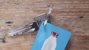 A set of keys and a blue tag with a milk bottle illustration on a wooden surface. - 'Het Melkhuis', the tiny house that makes you happy😀 (Katwijk aan Zee)