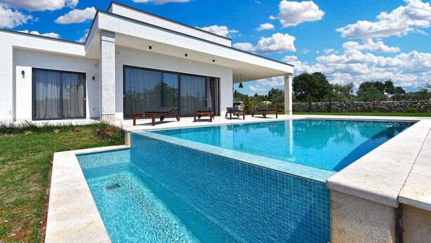 Ein modernes Haus mit einem großen Schwimmbad, einer überdachten Terrasse mit Liegestühlen und einer Steinmauer im Hintergrund.