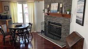 A dining area with a round table and chairs, a stone fireplace with a mantel, and a view of the outdoors through a window with curtains. - Cabana Too Smith Mountain Lake (Moneta)