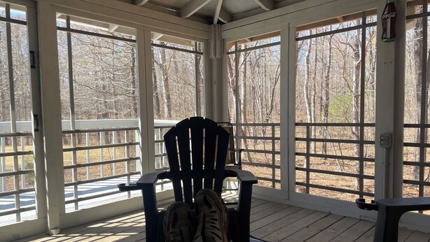 A covered porch with a wooden floor, a black chair, and a view of leafless trees through large windows. - Cabana Too Smith Mountain Lake (Moneta)
