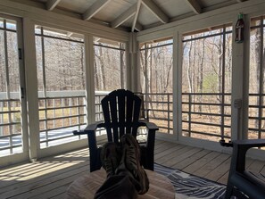 A covered porch with a wooden floor, a black chair, and a view of leafless trees through large windows. - Cabana Too Smith Mountain Lake (Moneta)