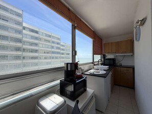 A compact kitchen with a view of a building across the window, a microwave, a kettle, and a small trash bin.