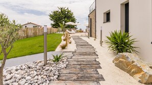 A pathway leading to a modern building with a stone facade, a wooden deck, and a well-maintained lawn. - Gîtes with heated pool near beach. Garden, spa, BBQ, WiFi. Baby bed included (Préfailles)