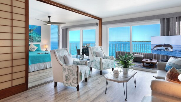 A spacious living area with a balcony overlooking the sea, featuring a sofa, armchairs, a coffee table, and a TV displaying a whale. - Alii Villas #332 Million Dollar Ocean View. Watch Dolphins, Coastal Vibes. (Kailua-Kona)